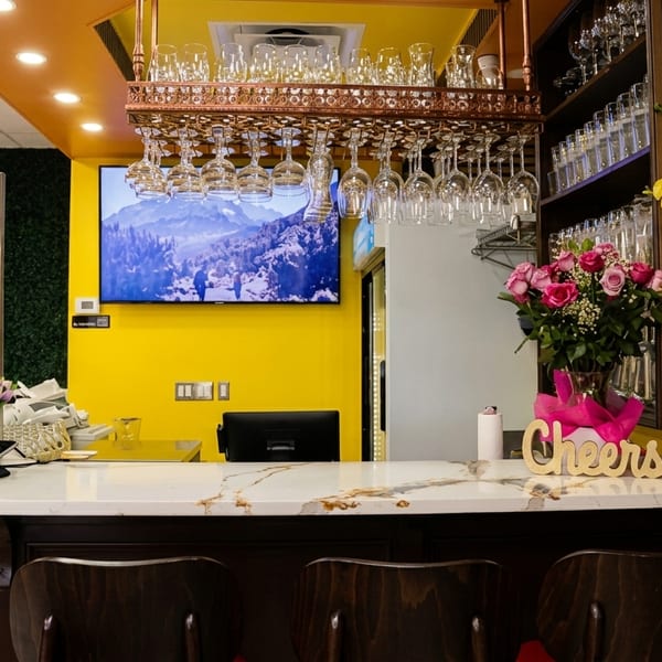 A vibrant yellow-walled restaurant interior with a pastry display, bar seating, and floral decorations.
