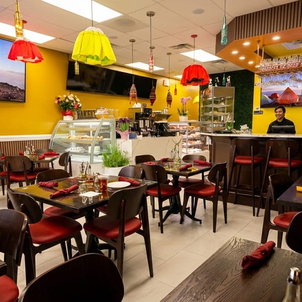 A brightly lit restaurant interior with yellow walls, dark wood tables, red chairs, and a smiling man behind the counter.