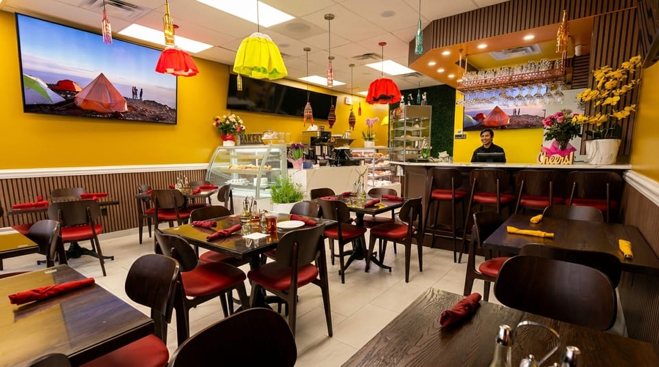 A brightly lit restaurant interior with yellow walls, dark wood tables, red chairs, and a smiling man behind the counter.