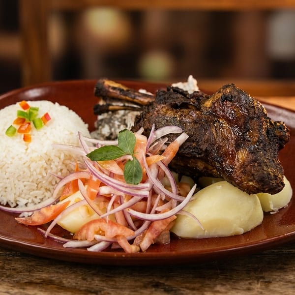A plate of roasted meat with rice, boiled potatoes, and a fresh red onion and tomato salad.